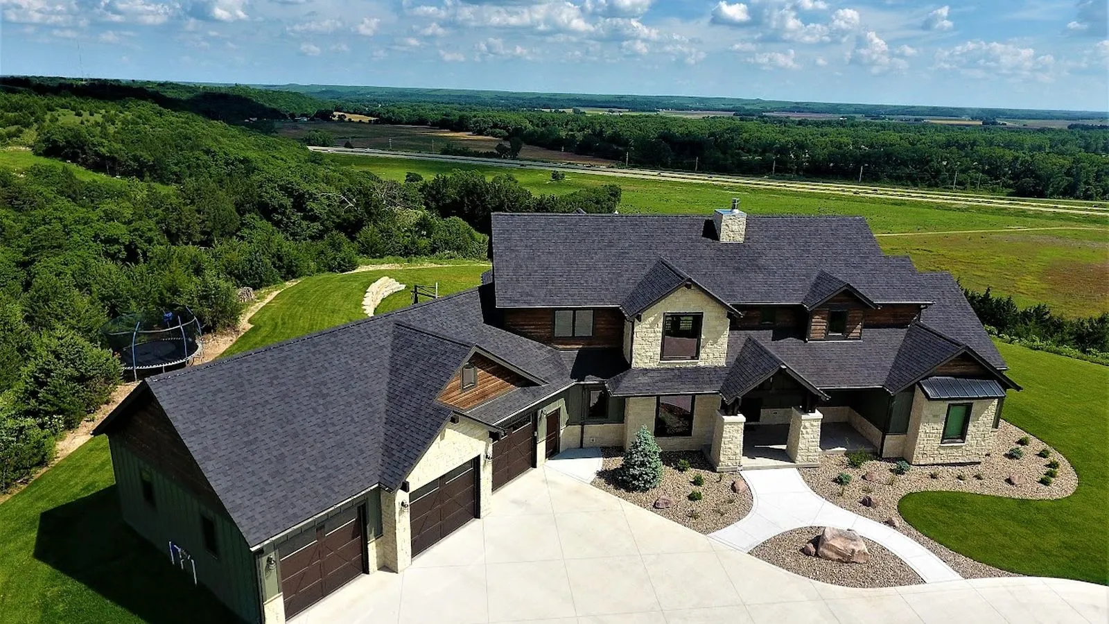 Aerial view of a stone and timber home with a new dark shingle roof overlooking rolling hills near Emporia, Kansas