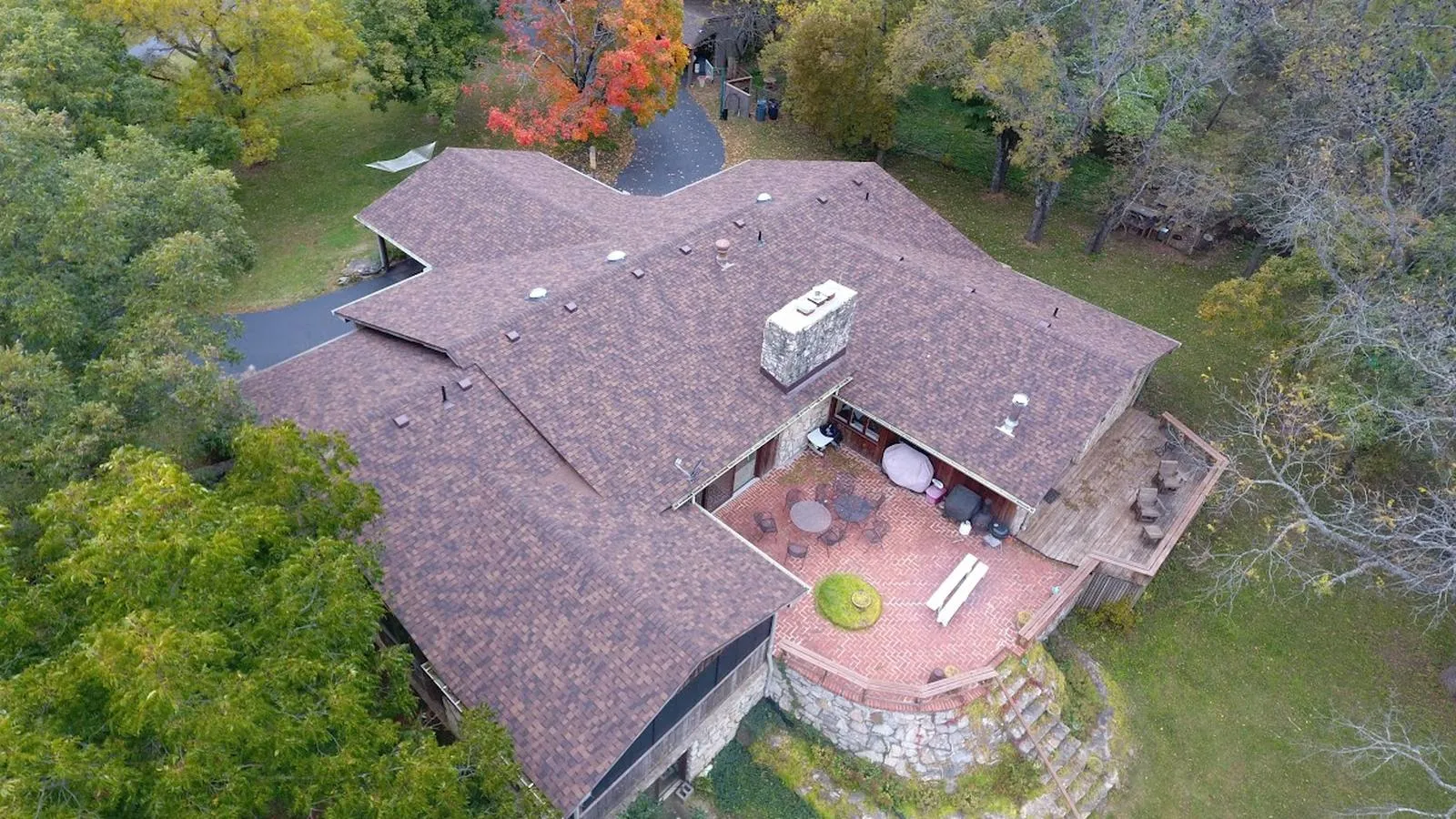 Aerial view of a stone Nebraska City home with a new brown architectural shingle roof, brick patio, and wooded grounds