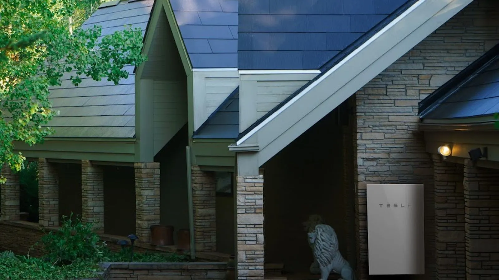 Stone home with a dark Tesla Solar Roof and a Tesla Powerwall battery mounted by the entry